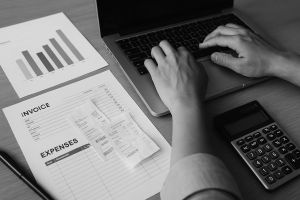 Black and white photo of hands typing on laptop with invoices and charts for advanced business accounting.